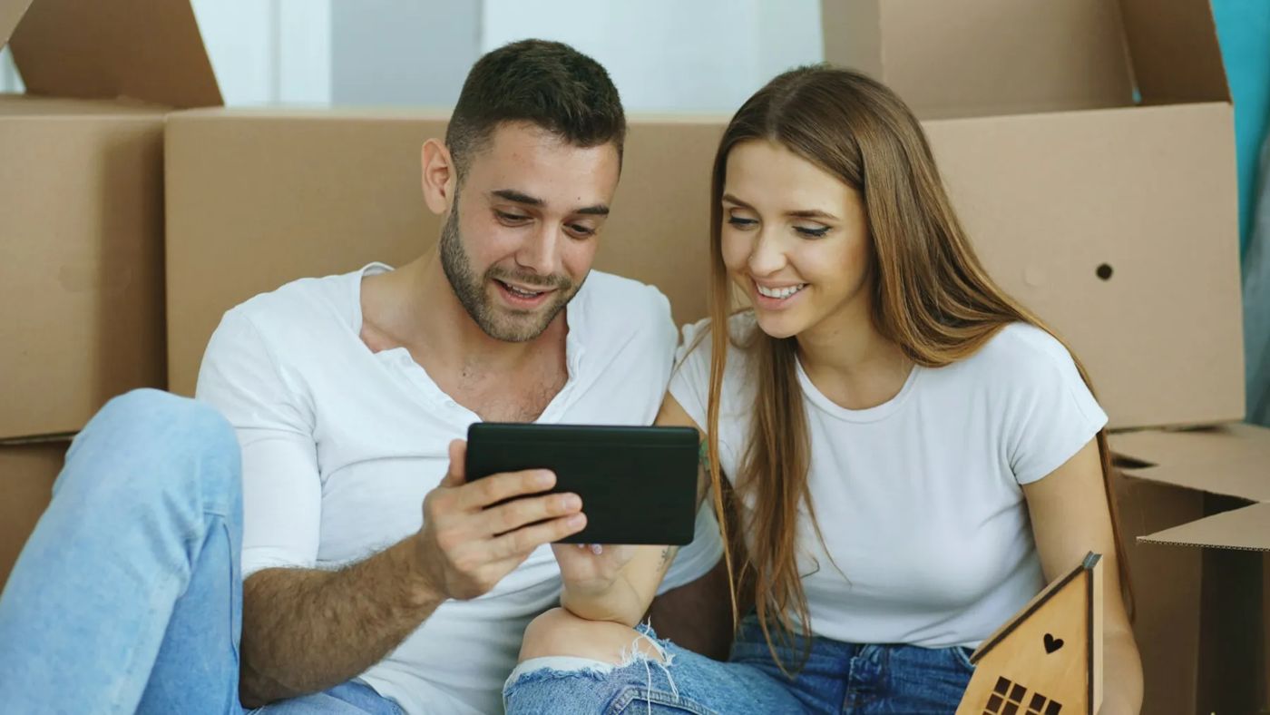 Junges Paar sitzt zwischen Umzugskartons in der neuen Wohnung und schaut auf ein Tablet
